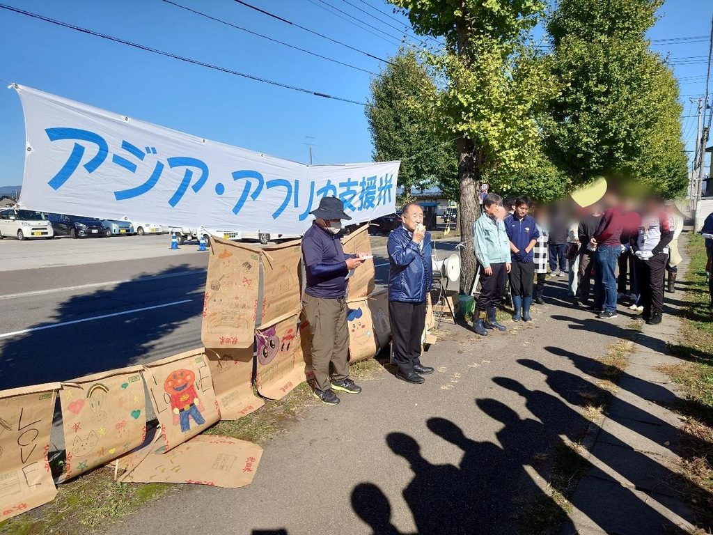 アジア・アフリカ支援米事業の主催者である石田寛さんが、横断幕の前でご挨拶をしている。インターアクト部がイラストを描いた米袋も見える。
